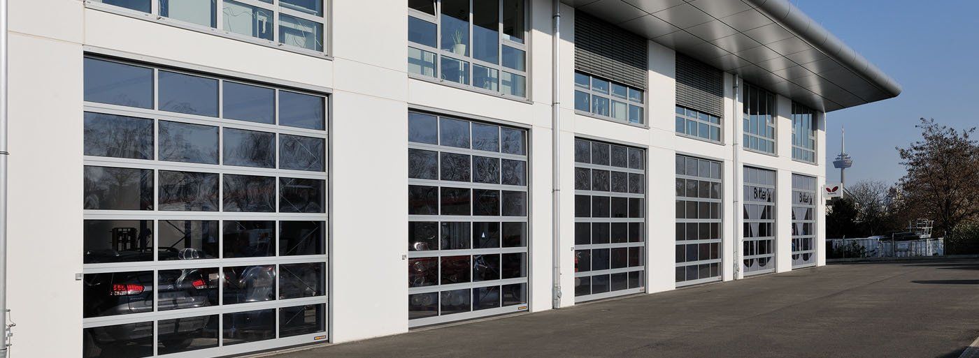 A white building with several large, clear glass garage doors. A car is visible inside.