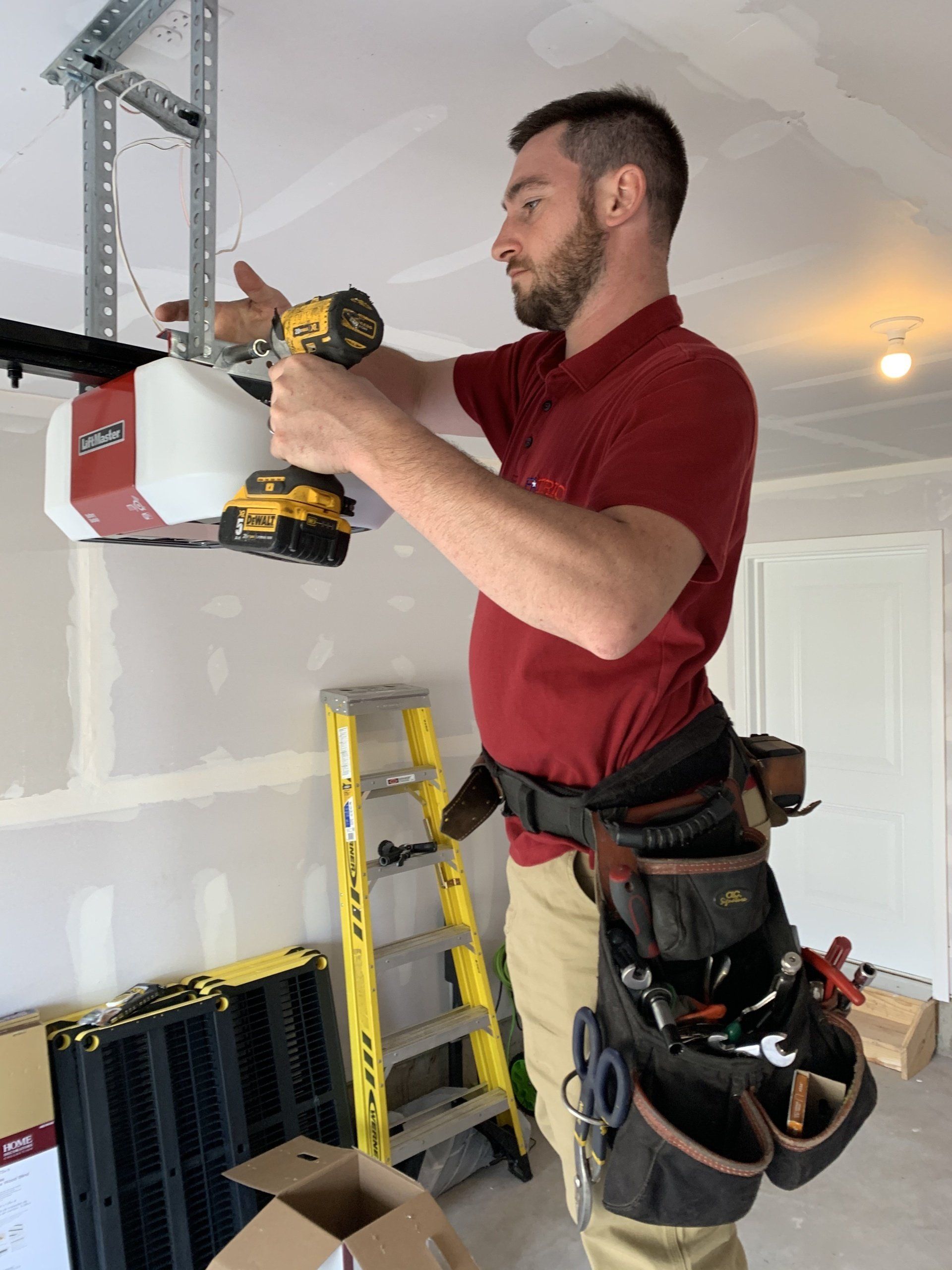 Man in red shirt installs a garage door opener, using a drill; he wears a tool belt, standing near a yellow ladder.