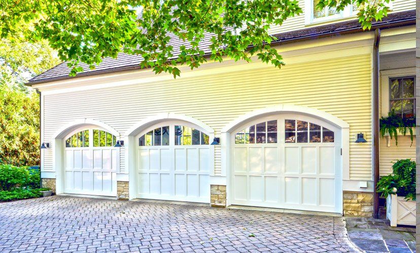 Three white garage doors with arched tops, on a light yellow house, cobblestone driveway, and green foliage.