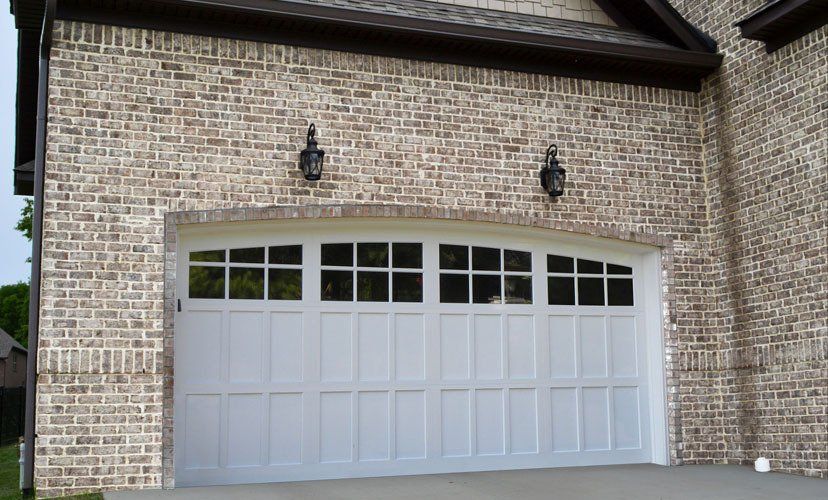 White garage door with upper windows, surrounded by brick wall, two lanterns above.