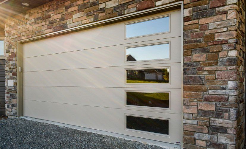 Beige garage door with four rectangular windows, set against a stone exterior wall.
