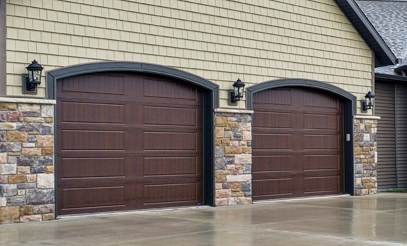 Two brown garage doors with arched frames, flanked by stone pillars, on a beige house.