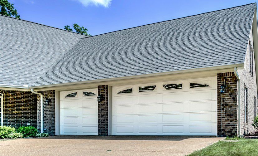 Two white garage doors with window panels under a grey roof, in front of a brick building.