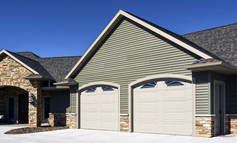 Two-car garage with beige doors, olive green siding, stone accents, and a dark roof against a blue sky.
