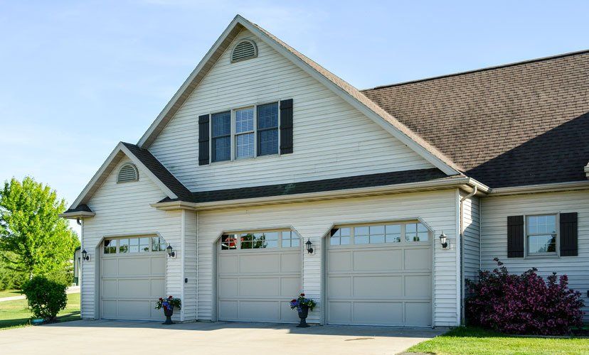 Three-car garage with light gray doors, white siding, and brown roof on a sunny day.