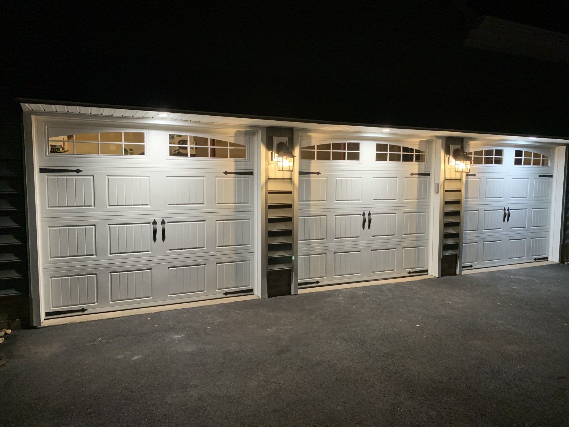Three white garage doors with windows and lights above, on an asphalt driveway.