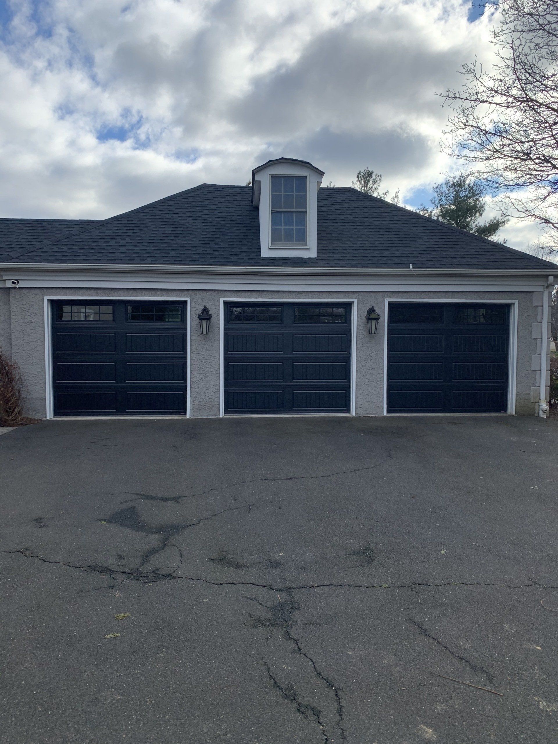 A three-car garage with black doors and a small window on a cloudy day.