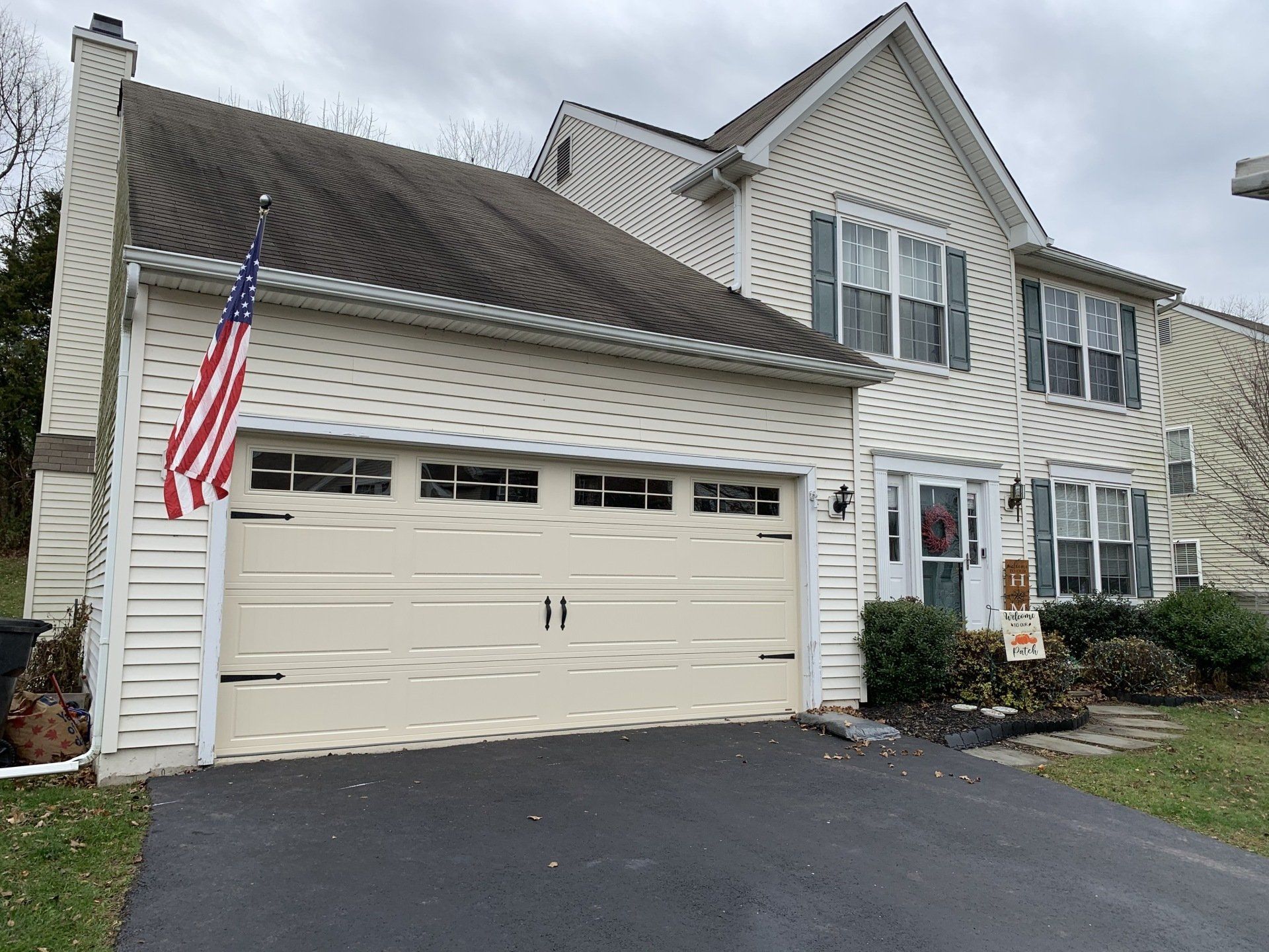 Beige house with two-car garage, American flag, and asphalt driveway on a cloudy day.