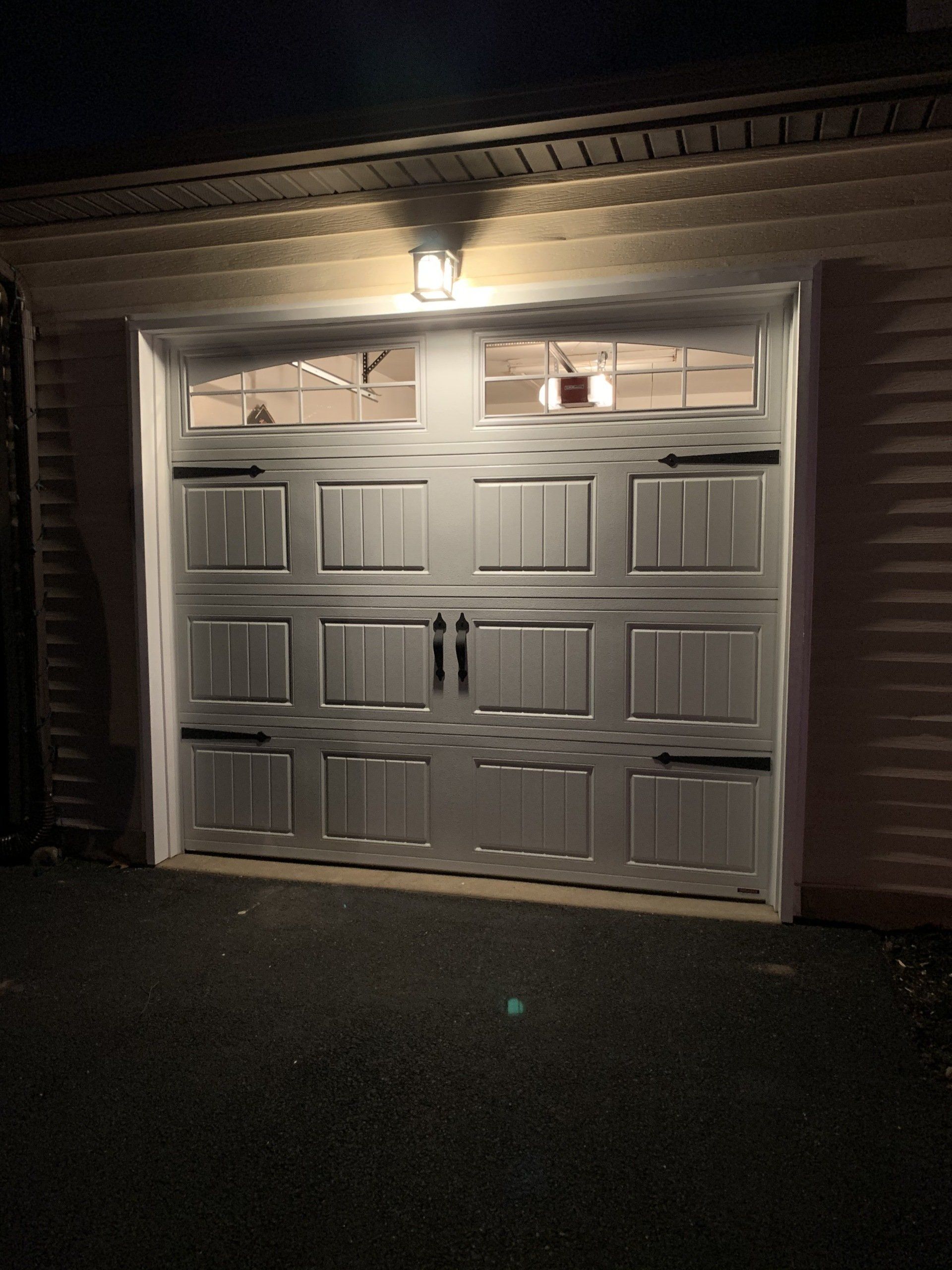 Lit garage door at night. Gray door with windows, black hardware, light above. Asphalt driveway.