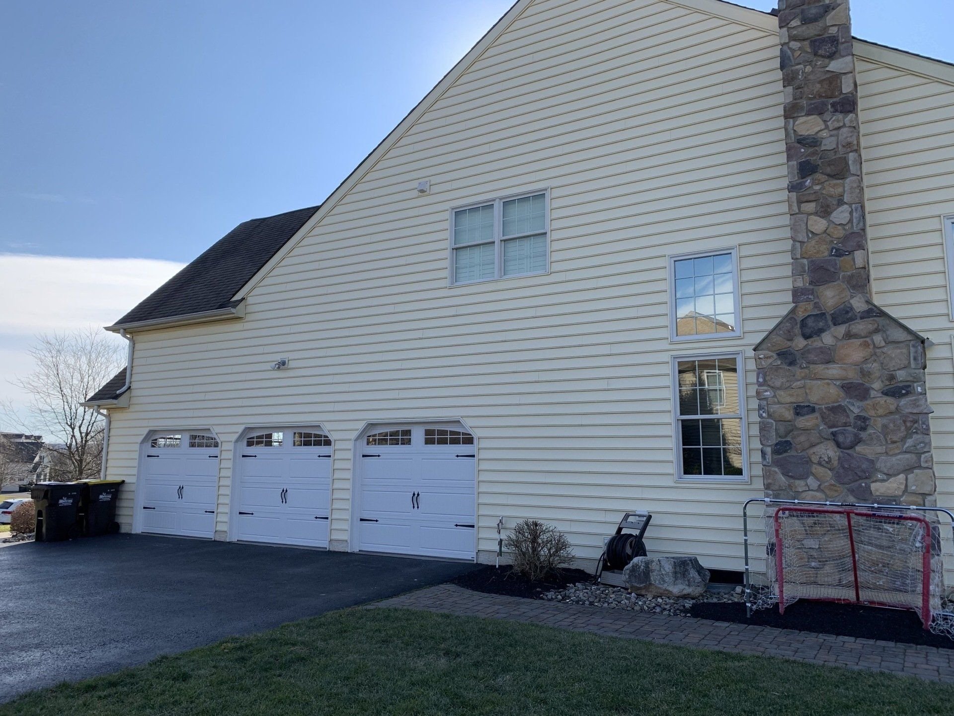 Cream-colored house with three white garage doors. Tall stone chimney, black driveway, green lawn, and a blue sky.
