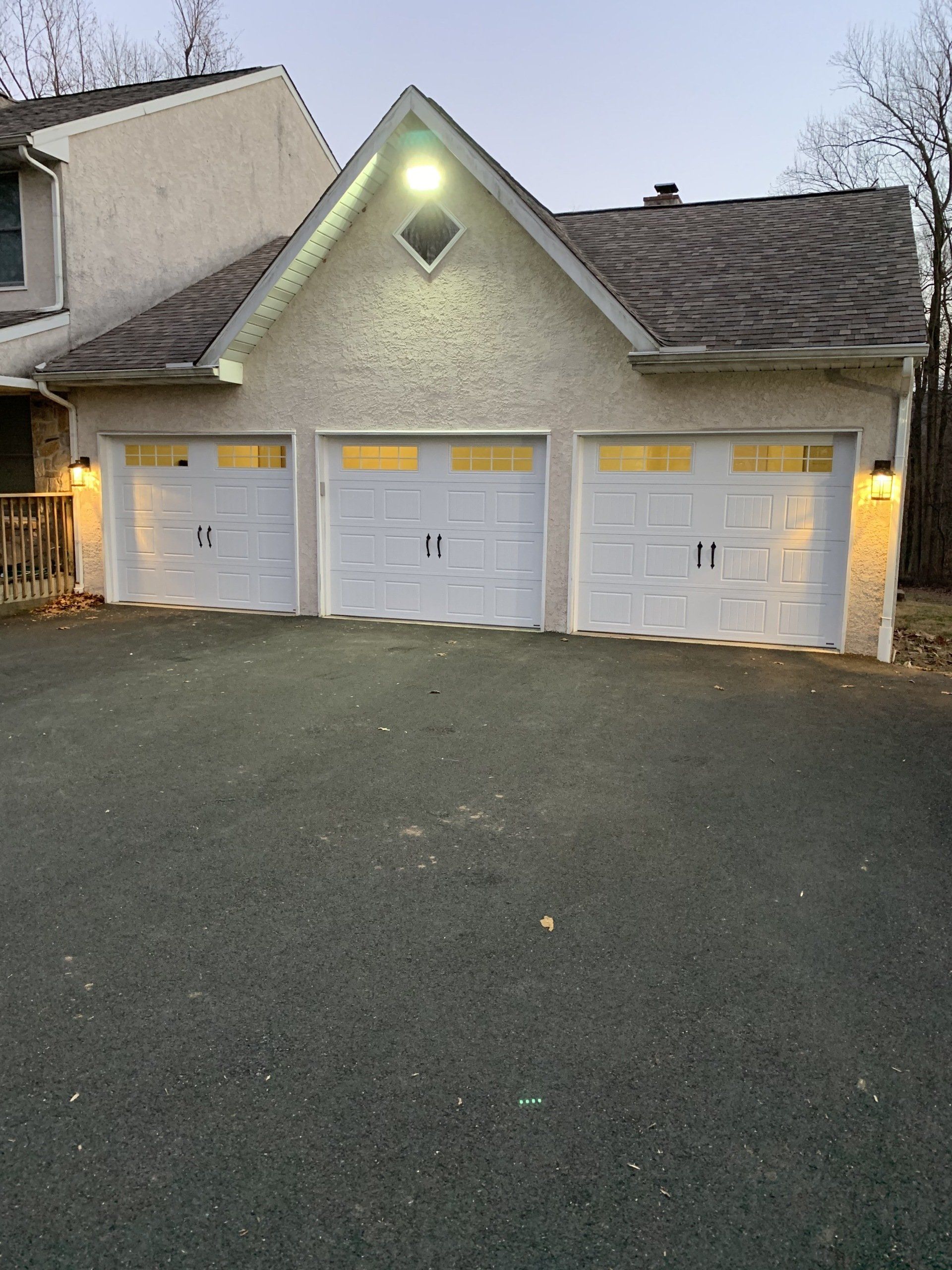 Three-car garage with white doors and tan stucco exterior; asphalt driveway; dusk lighting.