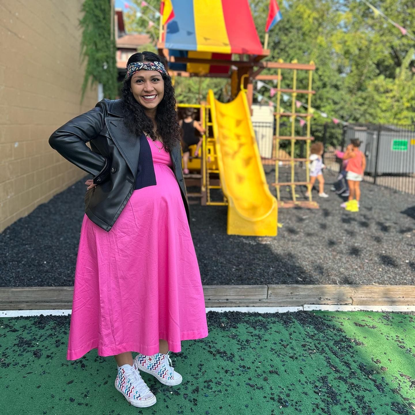 A pregnant woman in a pink dress is standing in front of a playground.