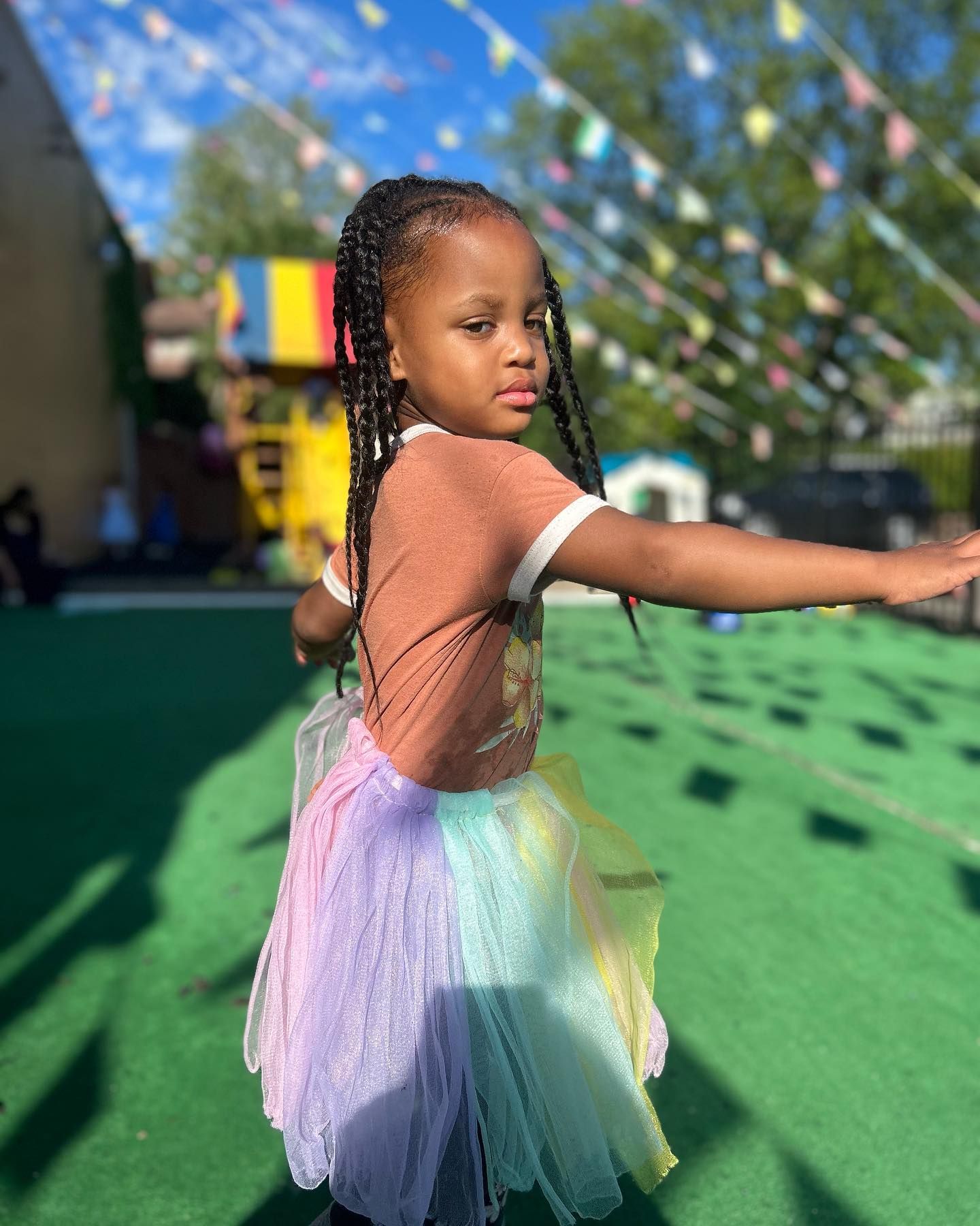 A little girl wearing a rainbow tutu is standing on a green field.