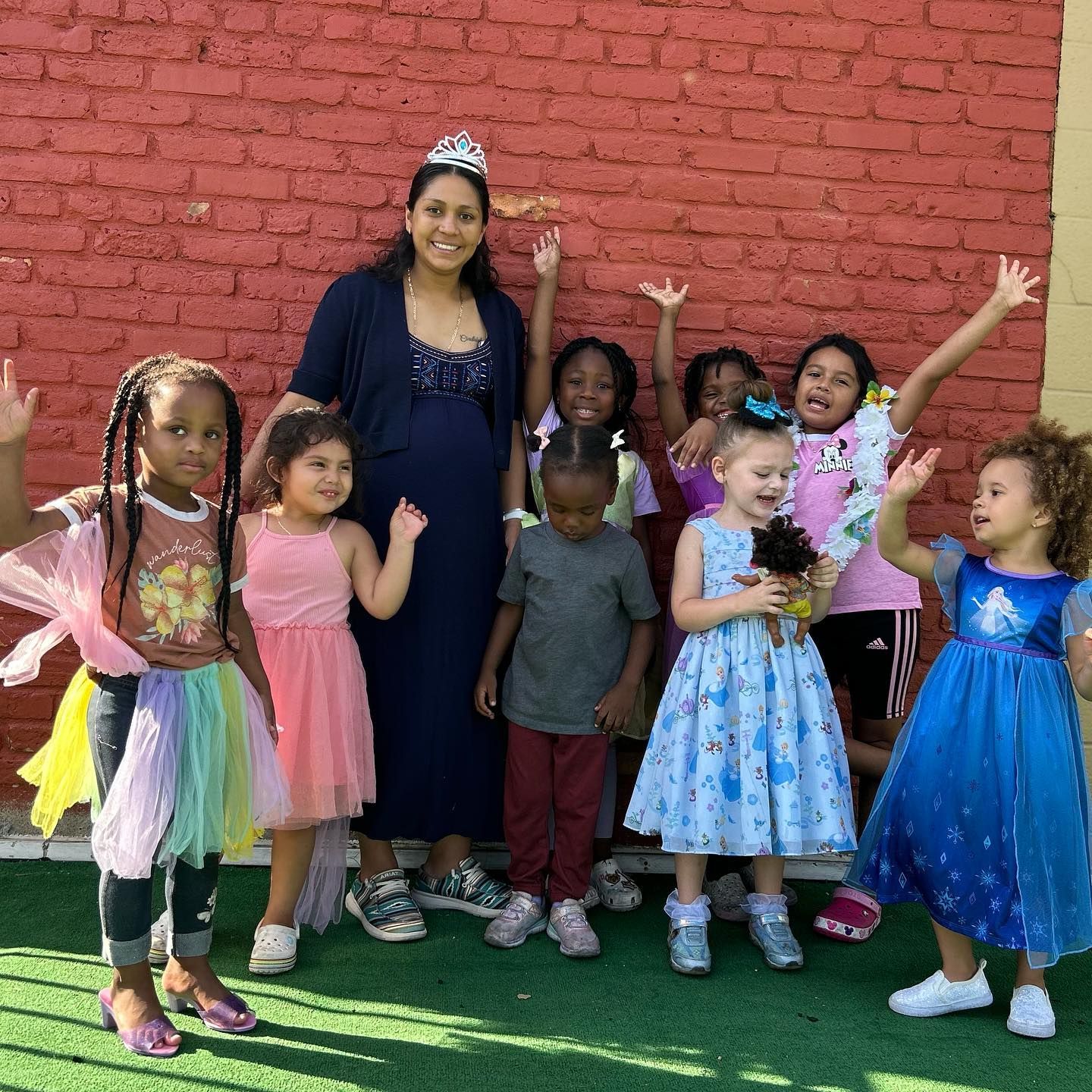 A woman with a tiara stands with a group of children, some in princess dresses, in front of a red brick wall.