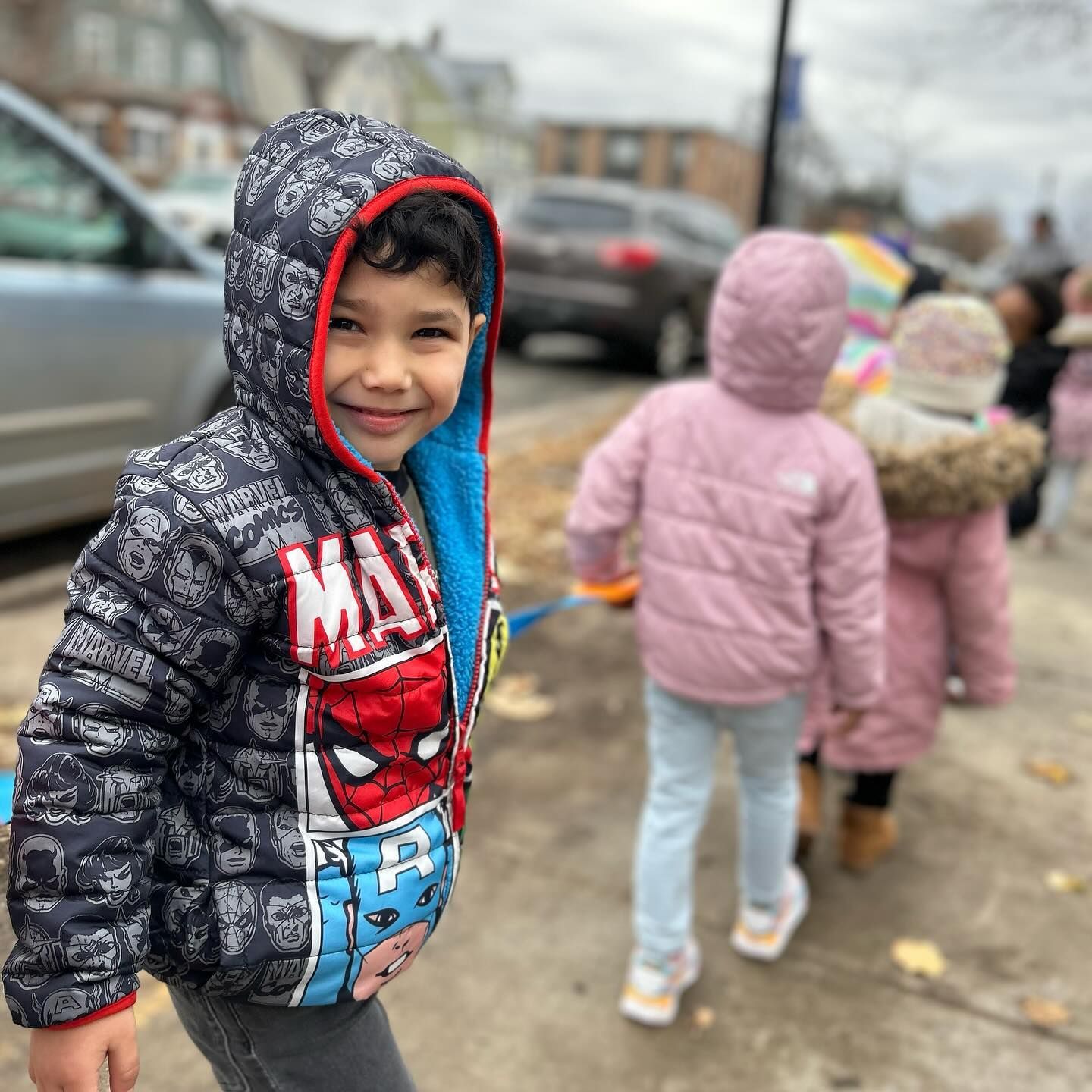 A young boy wearing a spiderman jacket is standing on a sidewalk.