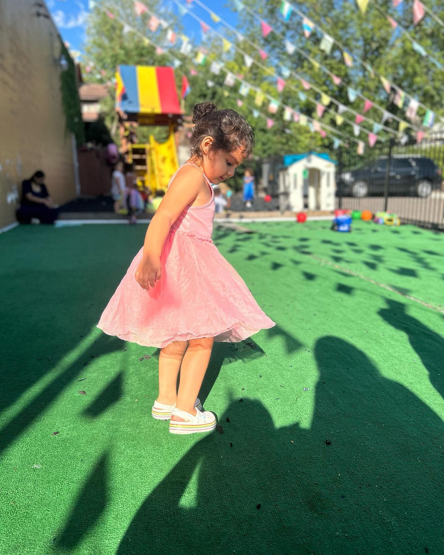 A young girl in a pink dress twirls on green turf outdoors, with a play structure and bunting in the background.