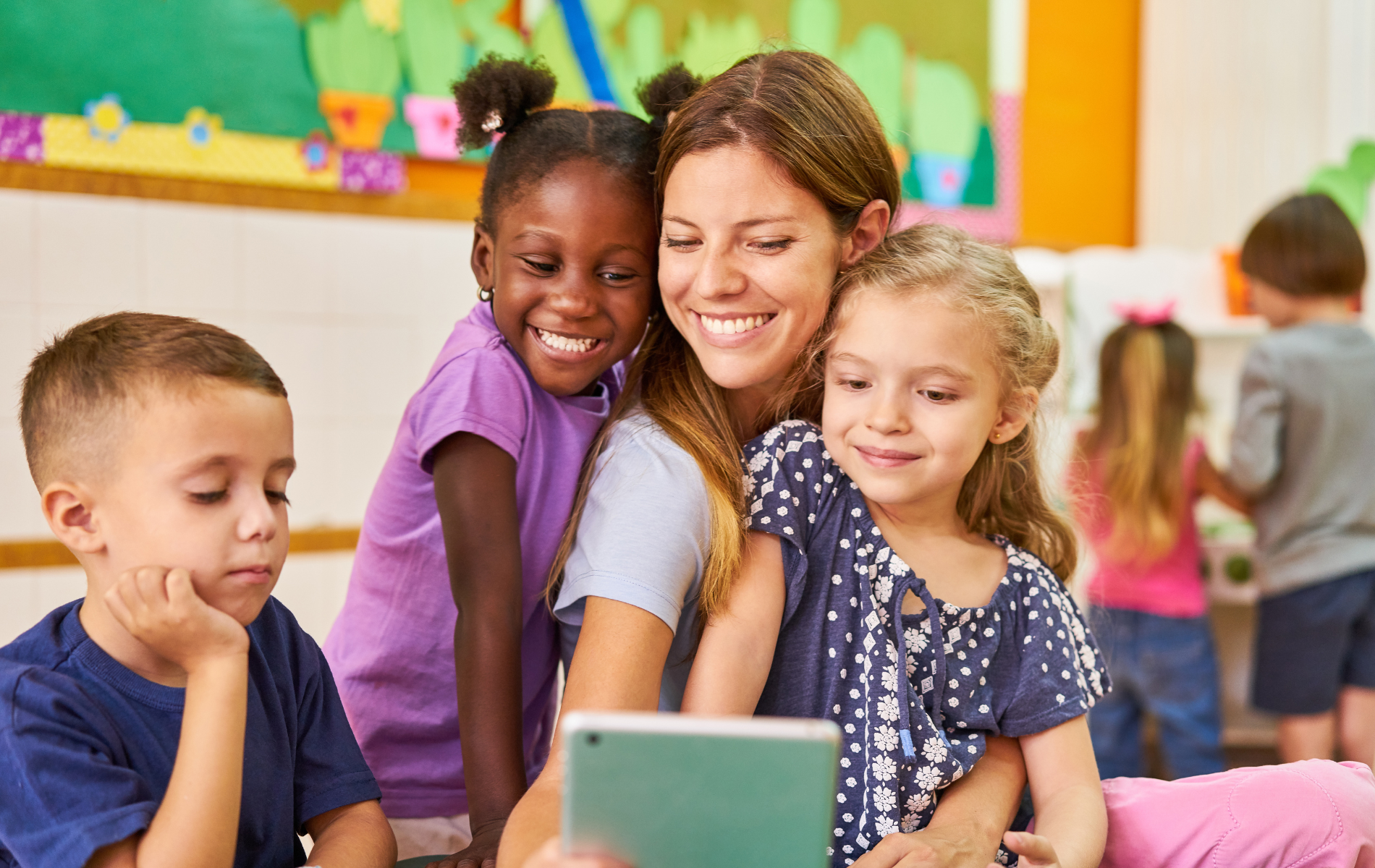 Teacher and children looking at tablet, smiling in a colorful classroom.