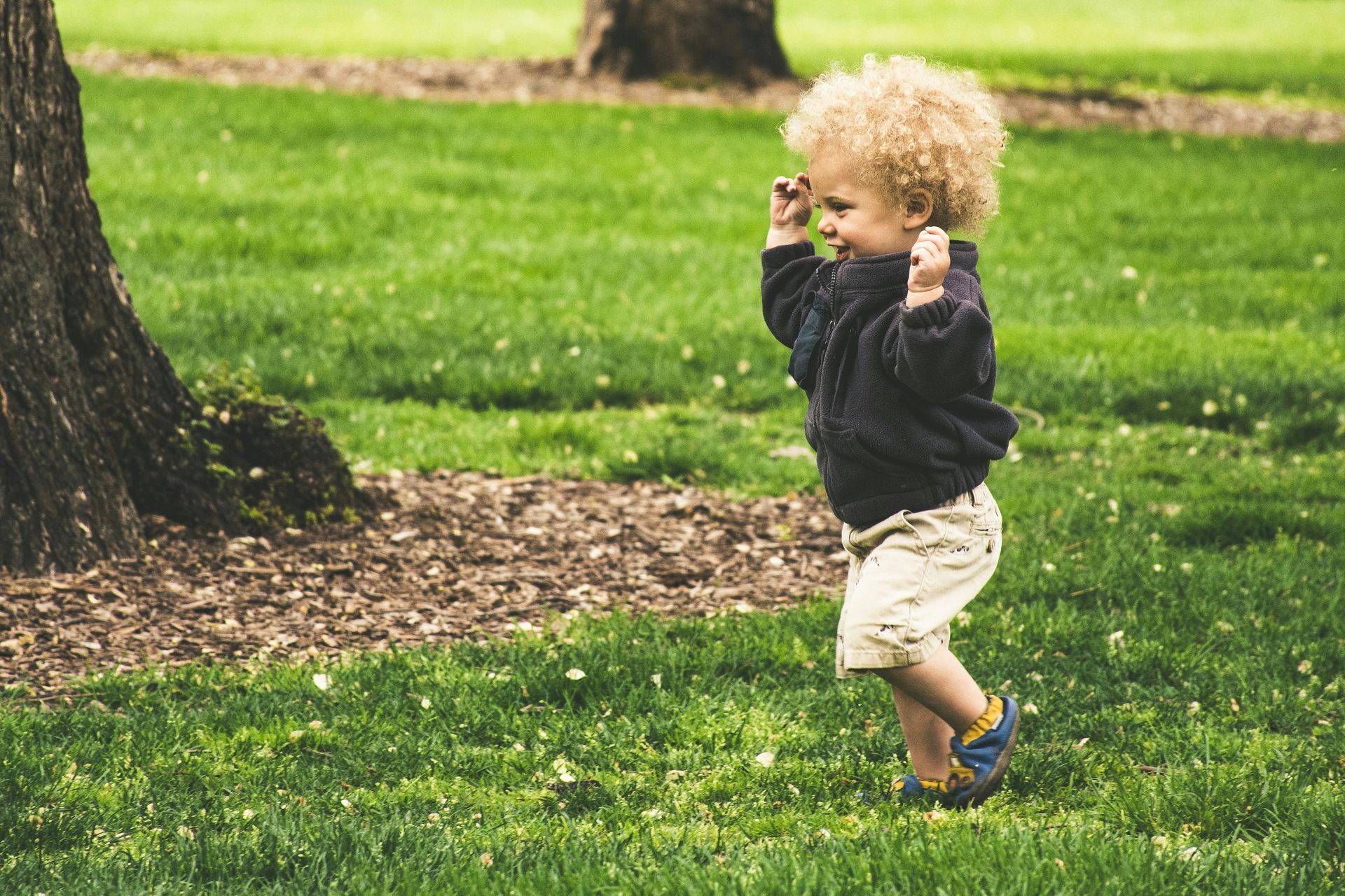 A young child with curly blond hair dances in a green park near a tree.
