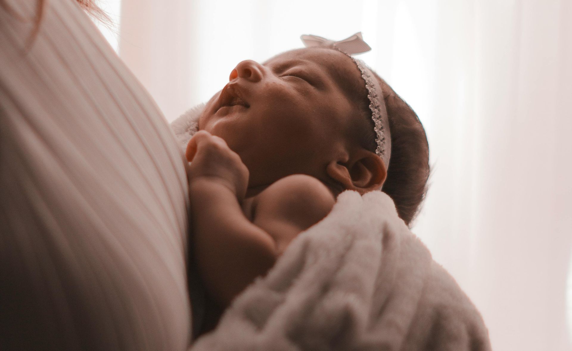 A newborn wearing a white headband with a bow rests against someone's shoulder, wrapped in a soft, light-colored blanket.