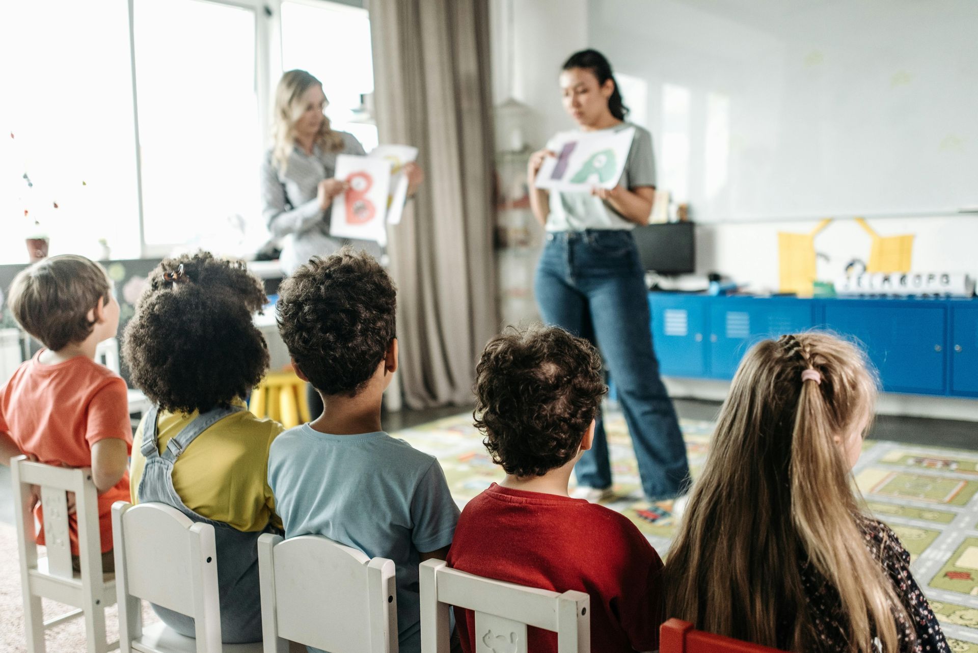 Children sitting in chairs, watching two teachers hold up alphabet letters in a classroom.