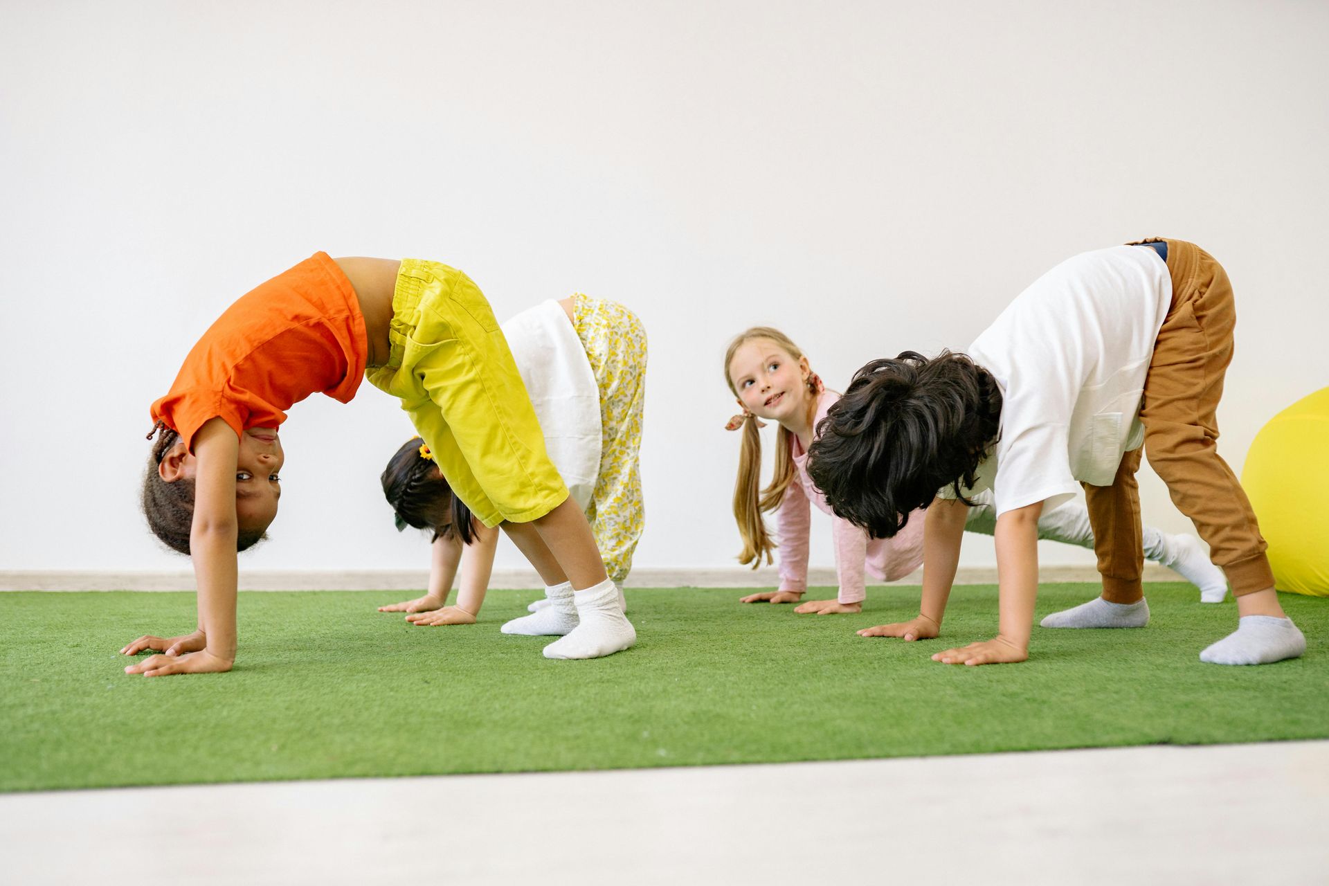 Children doing yoga, bending backward in a line on a green mat against a white wall.