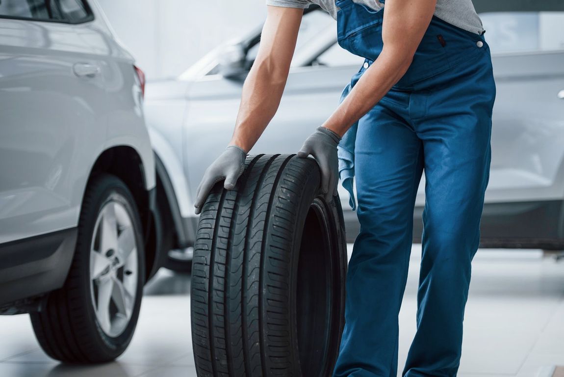 A man in blue overalls is holding a tire in a garage.