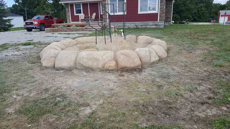 Round stone fire pit in a yard in front of a red house. A red truck is parked nearby.