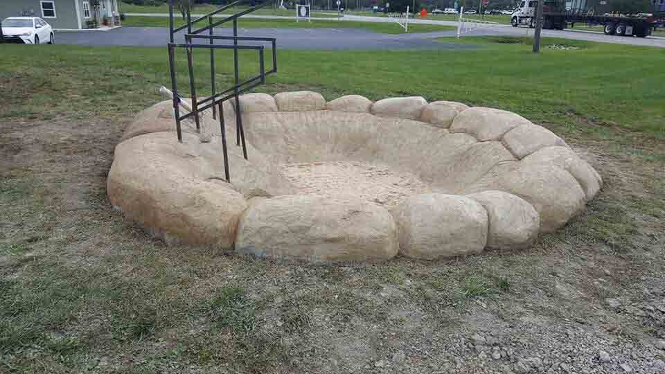Oval-shaped fire pit made of large stone-like structures, with a black metal structure rising from it, set on a grassy area.