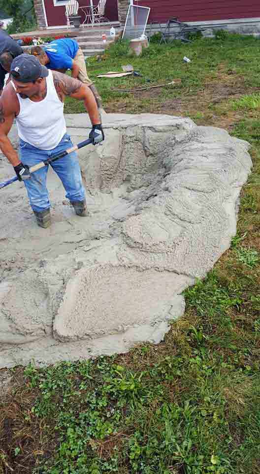 Man in white tank top using a shovel to smooth wet cement inside a pond-shaped structure, outdoors.