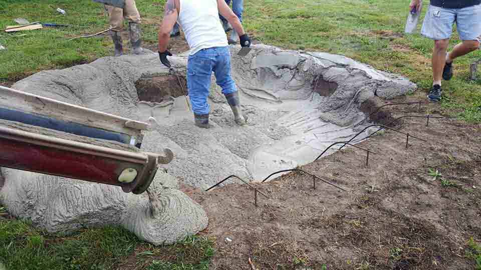 Workers pouring concrete into a pond form in a grassy area.