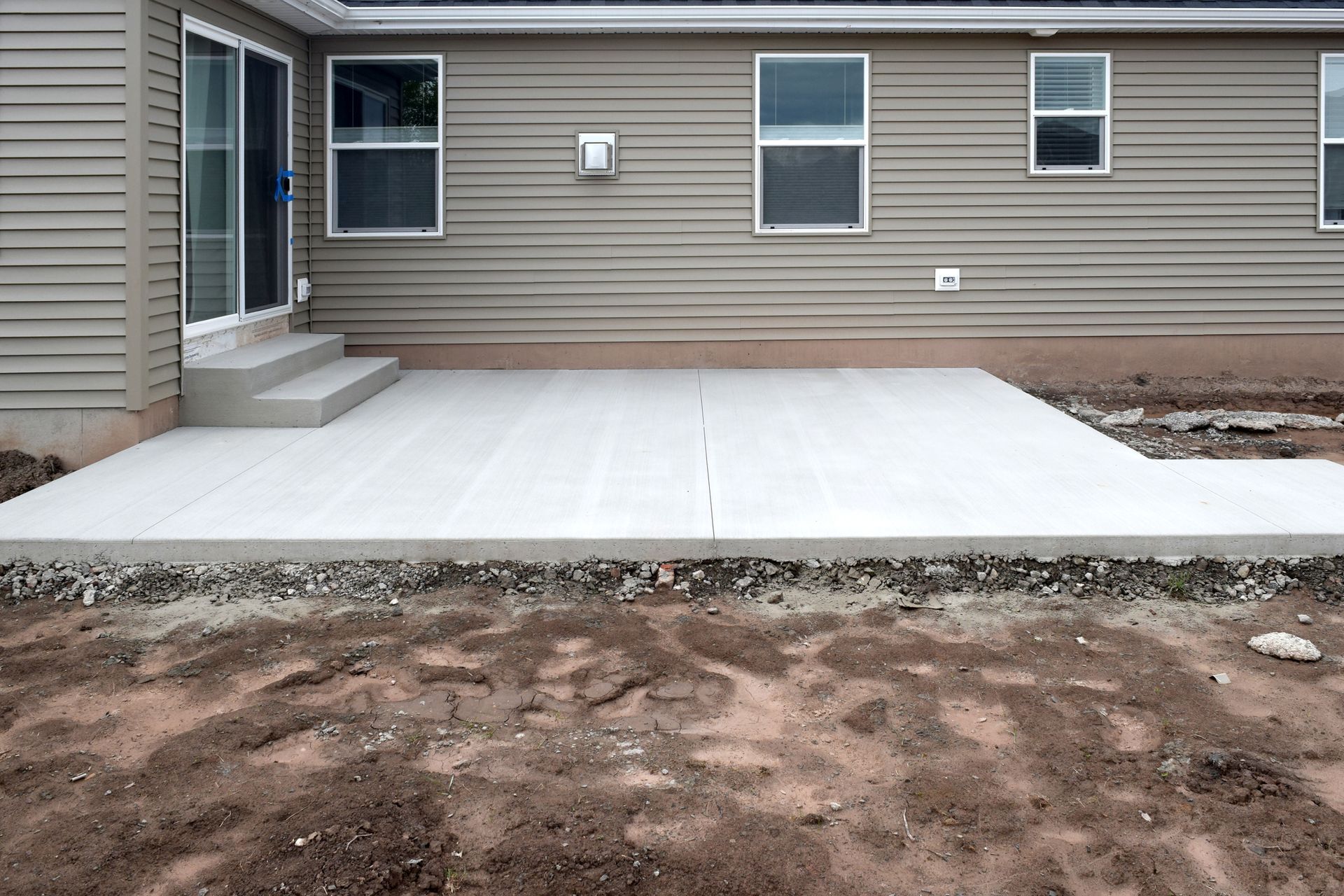 New concrete patio behind a house with steps leading to a sliding glass door and bare soil around.