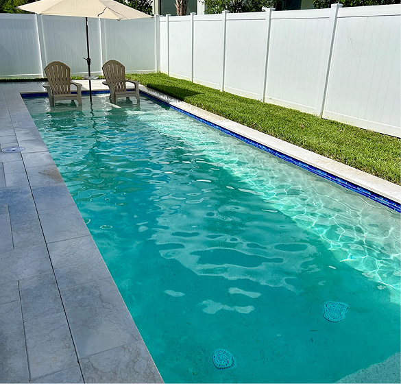 Rectangular pool with turquoise water, patio chairs, umbrella, and a white fence.