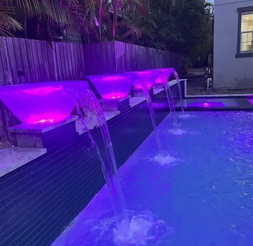 A nighttime pool with purple LED-lit waterfalls cascading into the blue water.