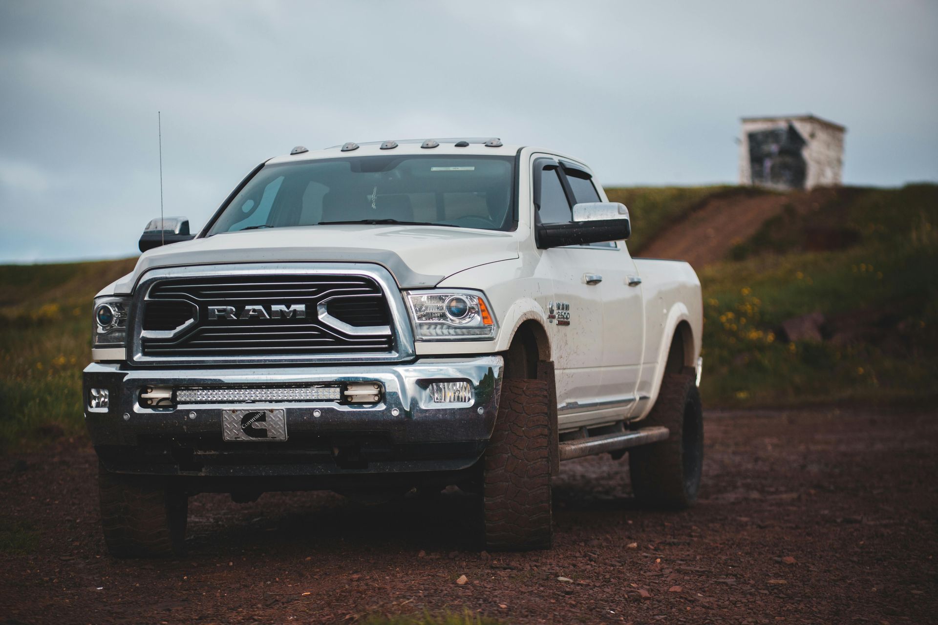 White RAM pickup truck parked on a dirt road under a cloudy sky with grassy hills behind it