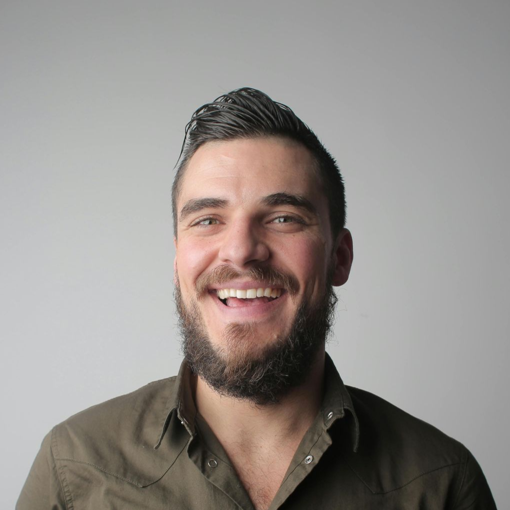 A person with a beard and short, styled hair smiles broadly against a plain gray background.