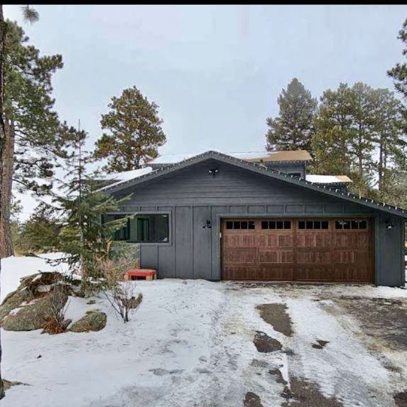 A house with a garage door is surrounded by snow and trees.