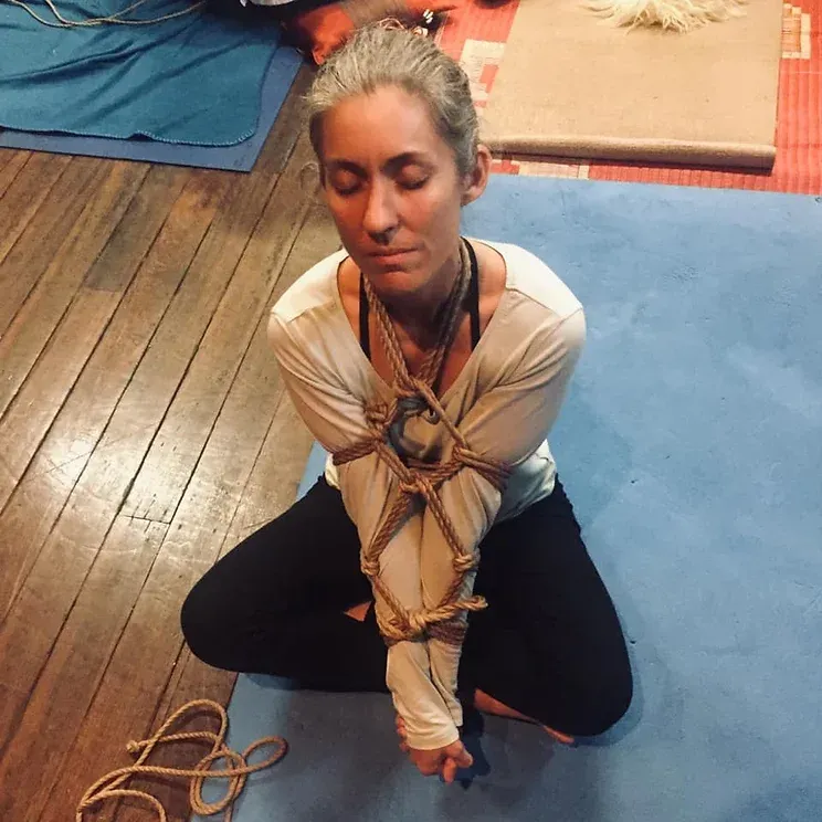 A woman is tied up and sitting on a yoga mat with her eyes closed