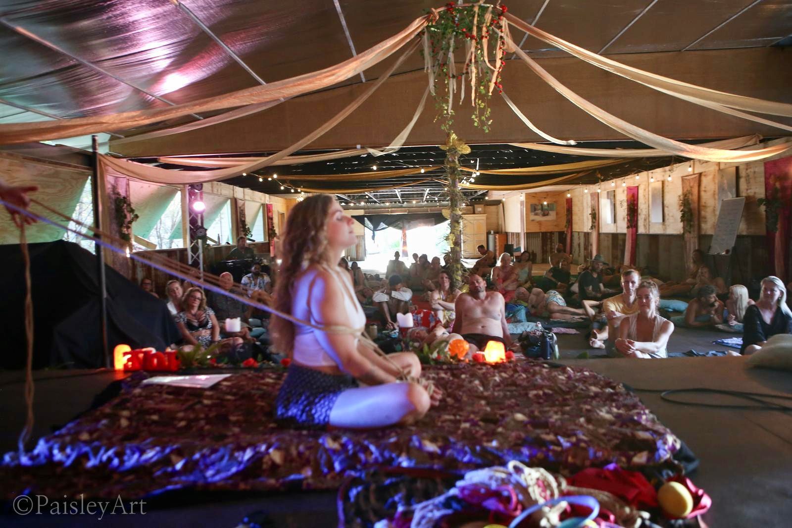 Shibari workshop at Taste Of Love Tantra Festival: A person sits in a meditative pose as she is tied in rope on a purple patterned mat under a canopy with fabric drapes in a crowded event space.
