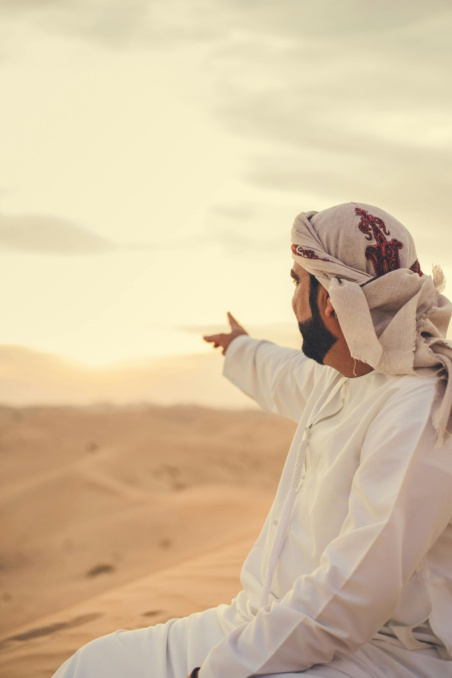 Man in traditional white clothing and head covering points towards desert horizon at sunset.