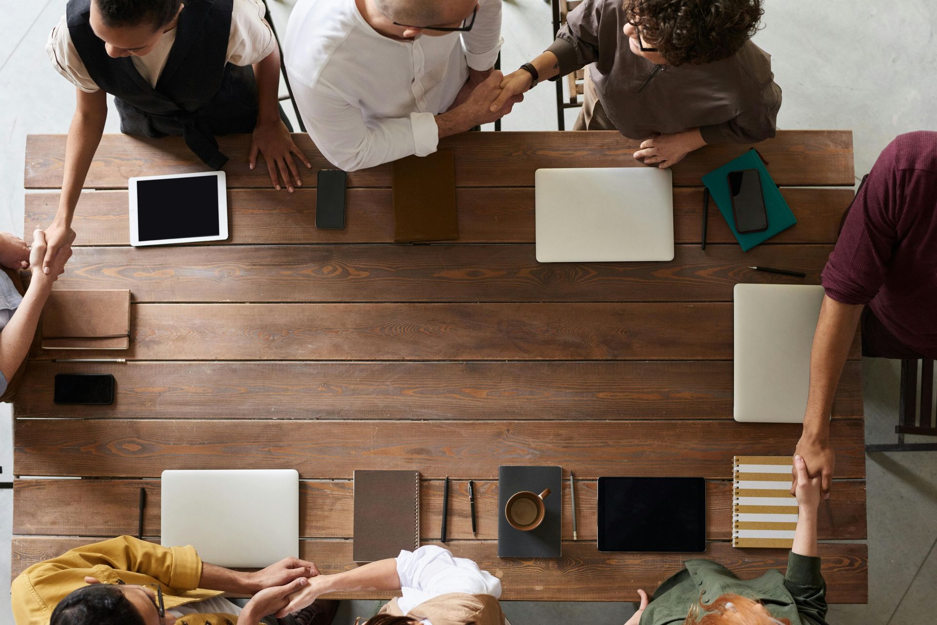 People shaking hands around a wooden table with laptops, phones, and notebooks.