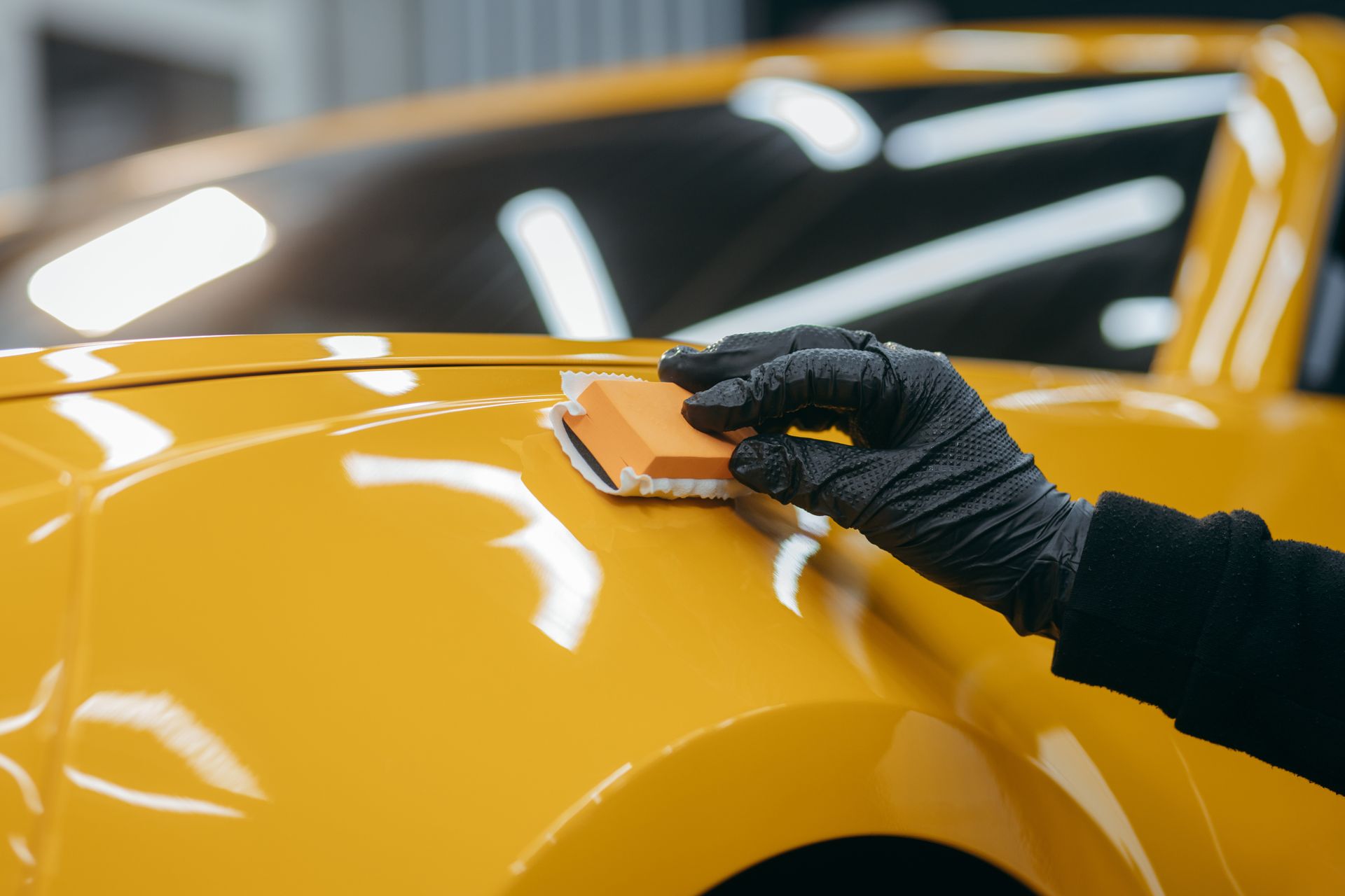 A gloved hand uses a small applicator block to apply a ceramic coating to the yellow hood of a car.