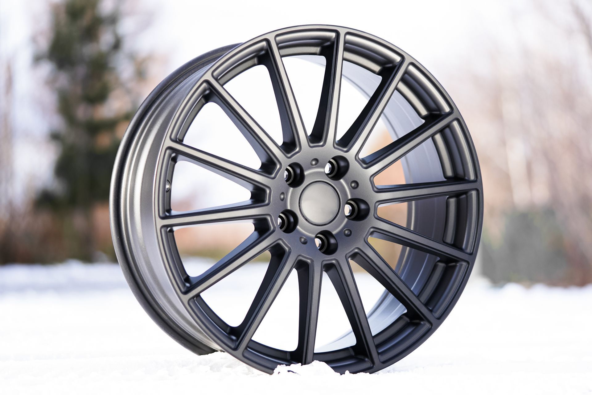 A dark gray, multi-spoke alloy wheel standing upright in a snowy, blurred outdoor setting.