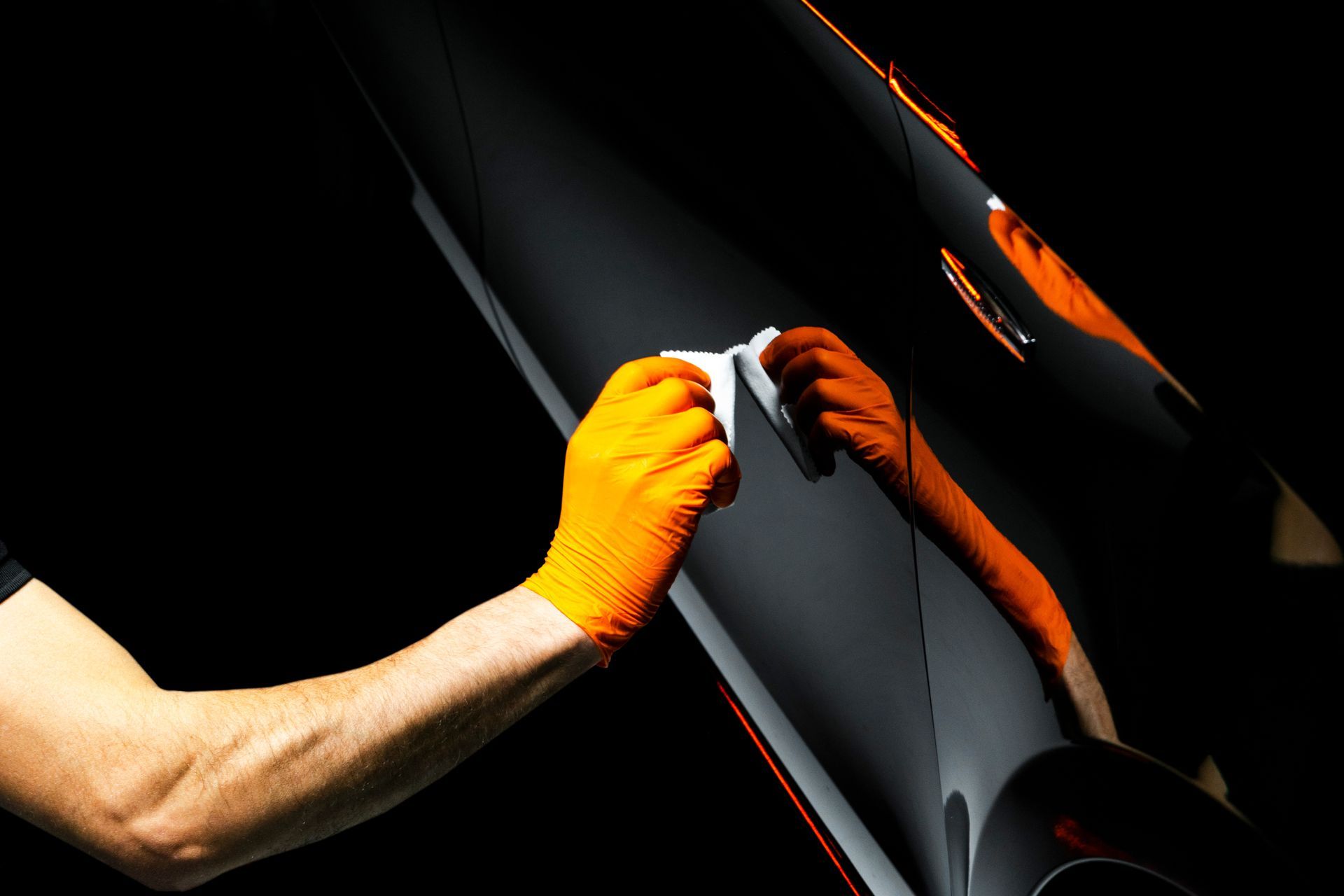 A hand in an orange glove wipes a shiny, black vehicle surface with a white cloth.