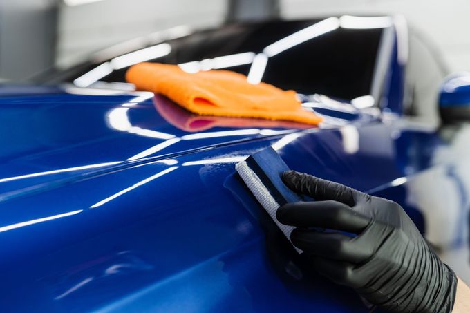 A gloved hand uses a foam block to apply ceramic coating to the glossy blue hood of a car, with an orange cloth nearby.