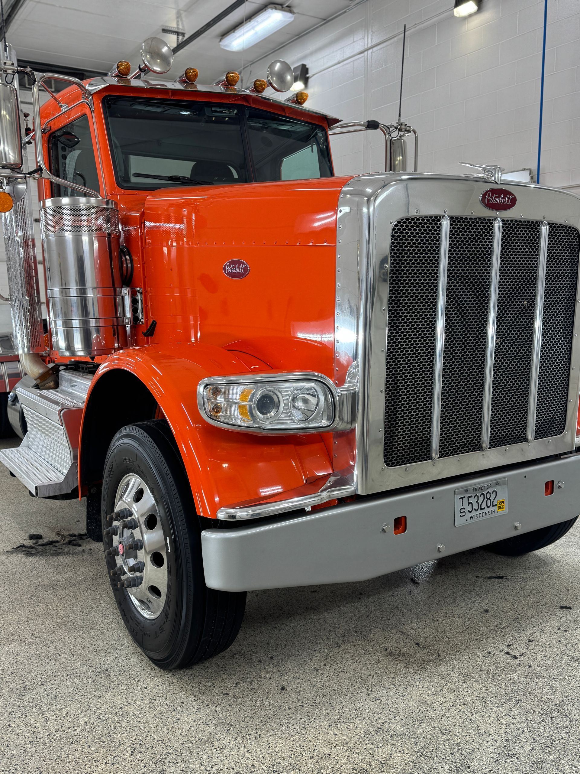 A bright orange Peterbilt semi-truck parked indoors on a speckled floor, featuring a prominent chrome grille.