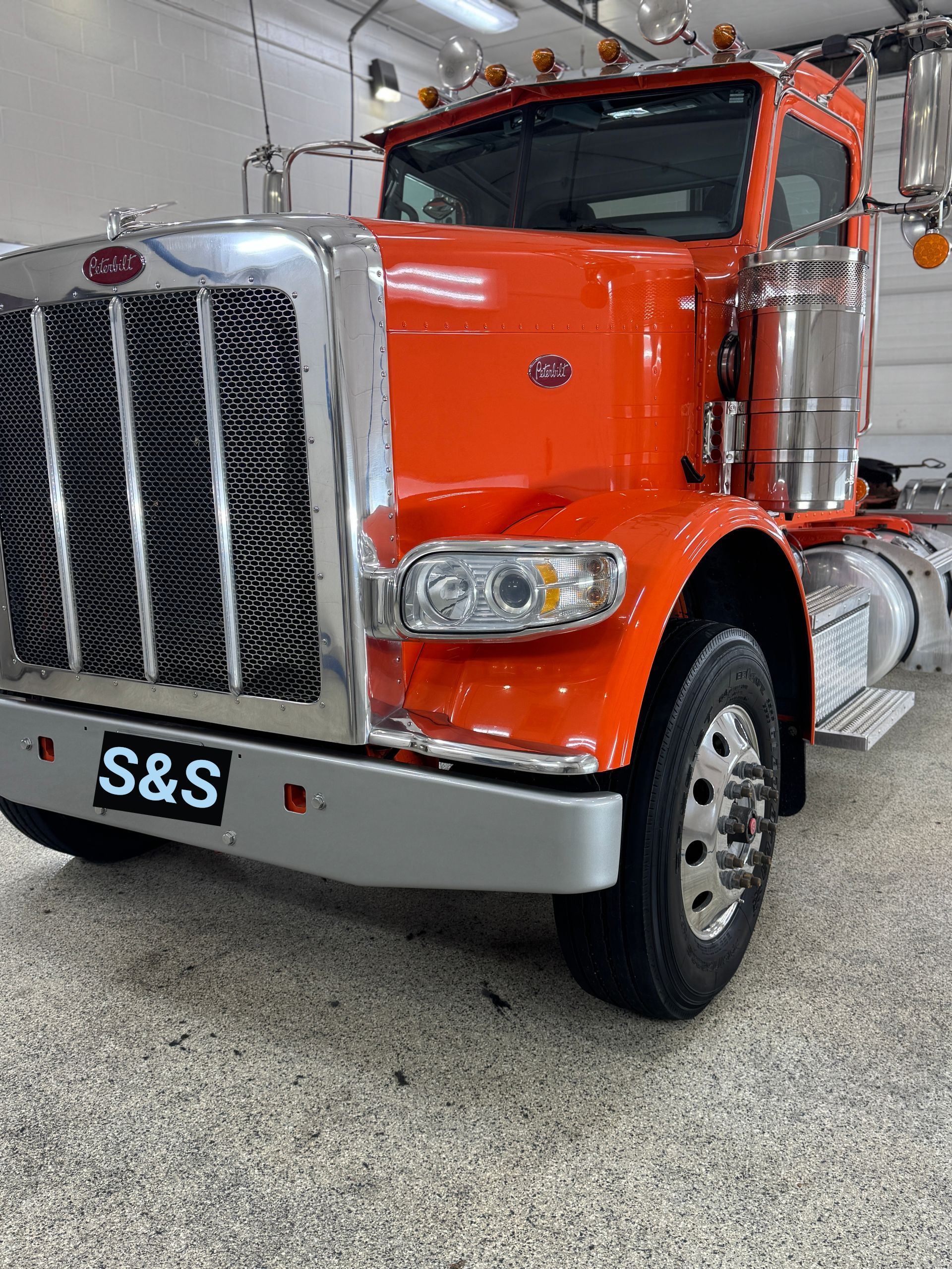 A bright orange semi-truck parked in a garage, featuring a large chrome grille and an S&S license plate on the bumper.