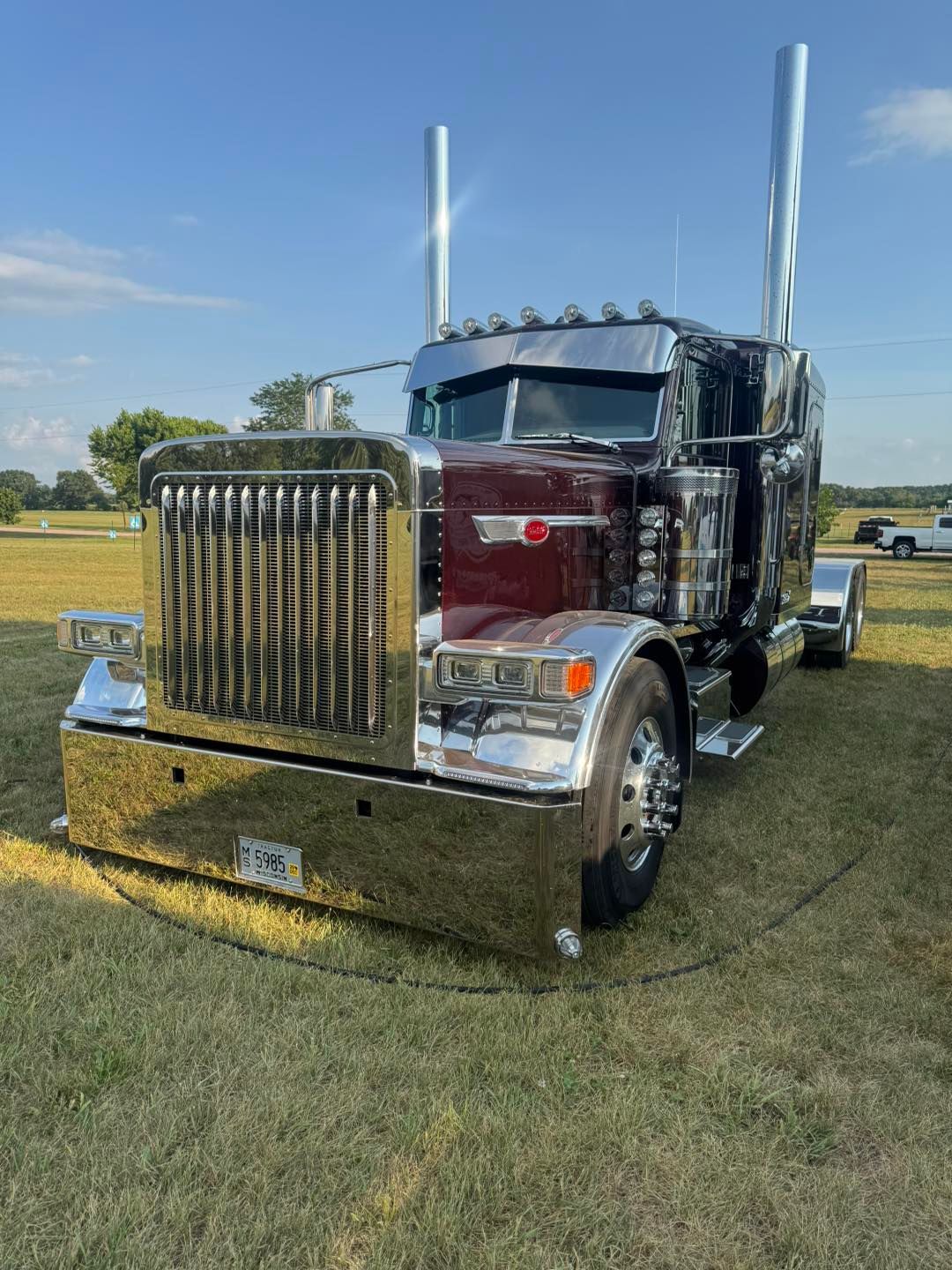 A customized maroon and chrome semi-truck parked on a grassy field under a clear blue sky.