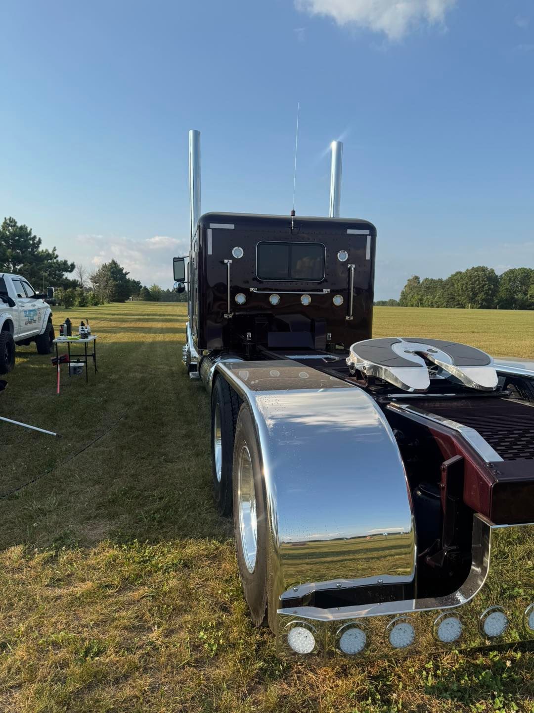 A dark-colored semi-truck with polished chrome fenders and exhaust stacks parked in a grassy field under a sunny sky.