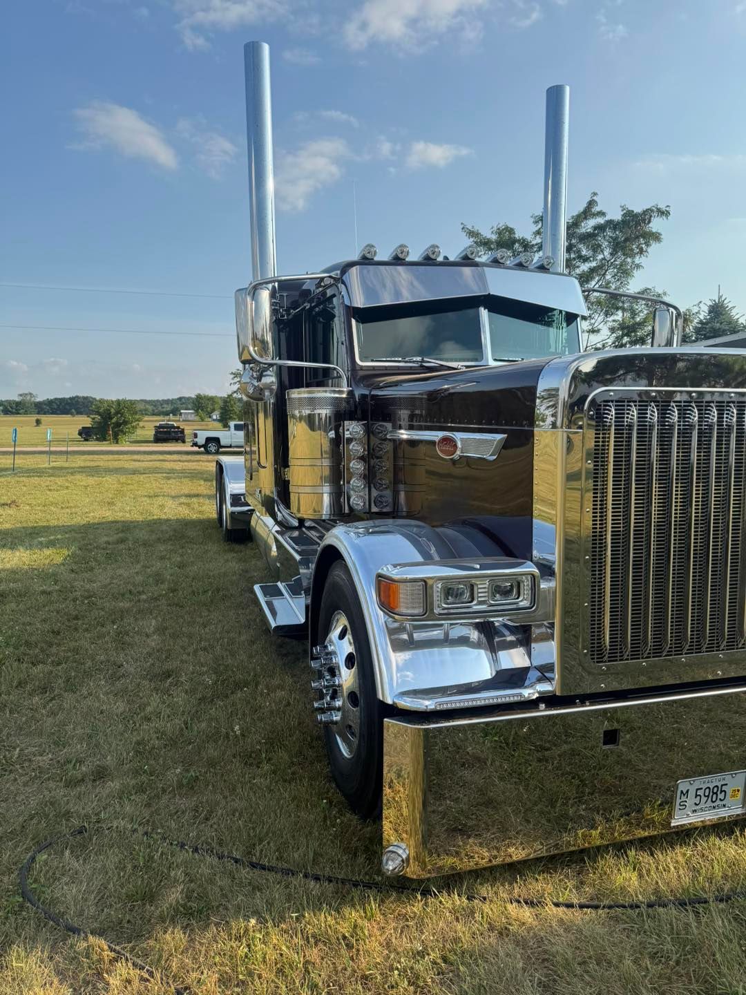 A polished brown semi-truck with large chrome exhaust stacks and a shiny bumper parked on a grassy field.
