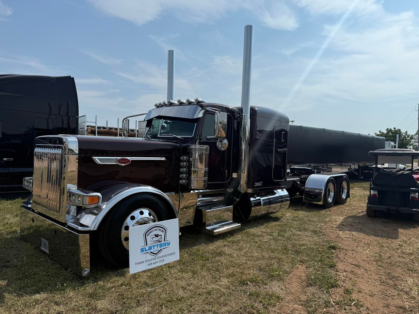 A dark maroon custom Peterbilt semi-truck with extensive chrome detailing parked in a grassy field on a sunny day.